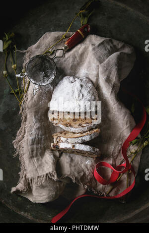 Tranches de gâteau de Noël traditionnel allemand, cuisson festive. Les stollen avec raisins secs et le sucre en poudre sur le linge de maison serviette avec tamis, ruban rouge, mis Banque D'Images