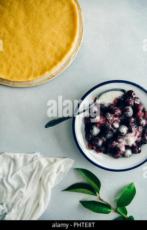 Cerises dénoyautées enrobées de sucre dans un bol blanc, pâte à tarte facile et feuilles vertes sur fond gris photographié en vue de dessus. Banque D'Images