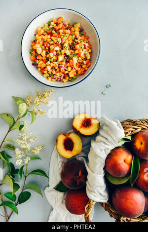 Salsa aux pêches dans un bol blanc accompagné d'un panier de pêches fraîches et fleurs minuscules photographié sur un fond gris en vue de dessus. Banque D'Images