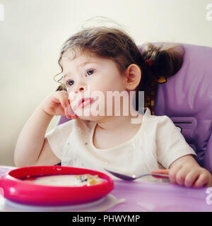 Portrait of cute ennuyer caucasien enfant jeune fille assise dans une chaise haute manger des céréales avec cuillère tôt le matin, la vie de tous les jours aux couleurs franches, moments Banque D'Images