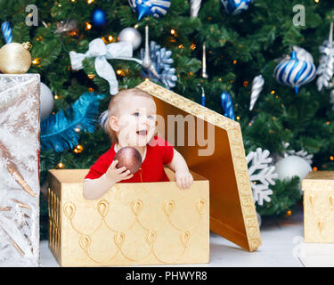 Portrait of happy funny smiling Caucasian baby girl toddler en rouge robe de vacances assis dans grand cadeau présent fort sous Nouvel An des arbres holding orname Banque D'Images