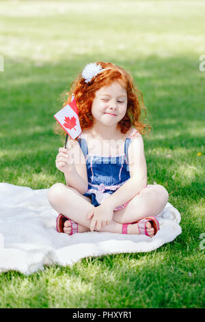 Portrait of cute little red-haired woman enfant tenant un drapeau canadien à feuille d'érable rouge sitting on grass in park à l'extérieur de la fête du Canada Banque D'Images