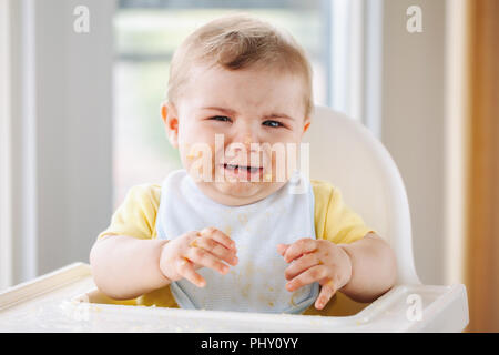 Portrait of cute pleurer caucasien enfant garçon avec désordre sale face sitting en chaise haute après avoir mangé de la purée de pomme avec les doigts. Accueil de tous les jours l'enfance Banque D'Images