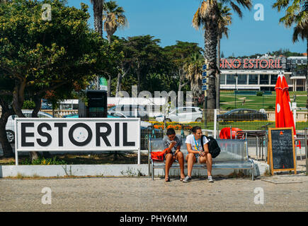 Estoril, Portugal - Août 30th, 2018 : deux adolescents s'asseoir sur un banc à côté d'un signe avec le géant casino Estoril dans dans le contexte Banque D'Images