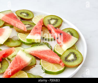 Plan des morceaux de pastèque, kiwi, citron vert et de la fonte des glaçons dans une assiette sur un fond de marbre gris avec de l'espace pour le texte Banque D'Images