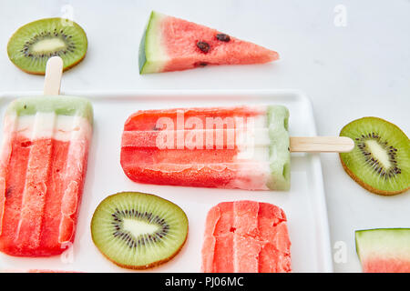 Watermelon ice cream on a stick présenté avec des morceaux de kiwi et juteux morceaux de melon dans une assiette sur un fond de marbre gris. Dessert d'été Banque D'Images