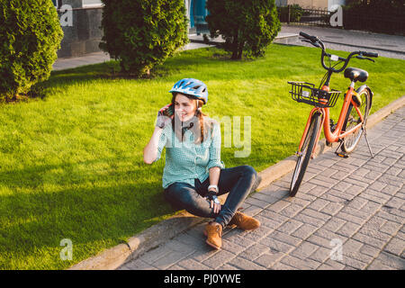 L'homme et de matériel roulant, vélo ville transports respectueux de l'environnement. Belle jeune femme de race blanche de repos assis sur l'herbe rouge utilise une mobil Banque D'Images