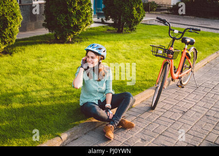 L'homme et de matériel roulant, vélo ville transports respectueux de l'environnement. Belle jeune femme de race blanche de repos assis sur l'herbe rouge utilise une mobil Banque D'Images