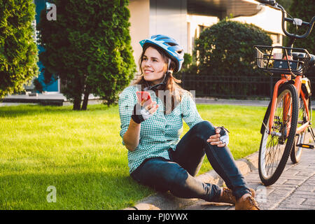 L'homme et de matériel roulant, vélo ville transports respectueux de l'environnement. Belle jeune femme de race blanche de repos assis sur l'herbe rouge utilise une mobil Banque D'Images