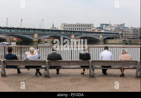 Une rangée de différentes personnes homme et femme assis sur des bancs à la recherche sur la tamise pendant leur pause déjeuner dans le centre de Londres. Banque D'Images