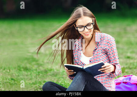 Portrait of a cute young brunette étudiant dans le parc ayant l'amusement et la lecture d'un livre Banque D'Images