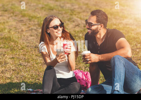 Pretty student girl à blanc T-shirt portant des lunettes de soleil et multiculturelle en étudiant-boy t-shirt noir et des lunettes de soleil assis sur l'herbe, boire du café et de parler les uns avec les autres Banque D'Images