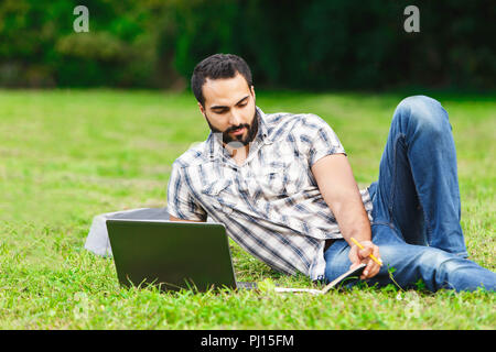 Un bel homme cheveux noir portant sur l'herbe et à la recherche sur son ordinateur portable. Il est à se détendre et profiter d'une journée ensoleillée Banque D'Images
