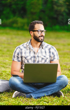 Jeune homme barbu en chemise à carreaux portant des lunettes assis sur l'herbe dans le parc avec son ordinateur portable et à la recherche autour de la belle journée ensoleillée. Banque D'Images