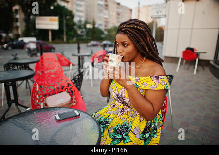 Hauteur petit mignon african american girl avec des dreadlocks, d'usure au niveau de la couleur jaune robe, assis au café en plein air sur la chaise rouge et boire du café. Banque D'Images