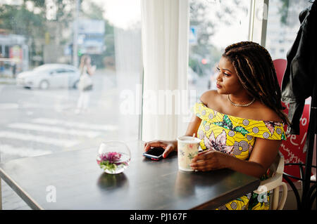 Hauteur petit mignon african american girl avec des dreadlocks, d'usure au niveau de la couleur jaune robe, assis au café avec tasse de café et en regardant la fenêtre. Banque D'Images