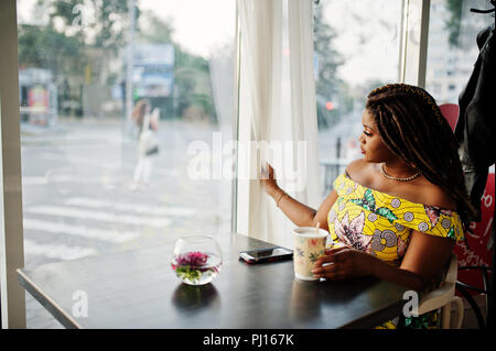 Hauteur petit mignon african american girl avec des dreadlocks, d'usure au niveau de la couleur jaune robe, assis au café avec tasse de café et en regardant la fenêtre. Banque D'Images