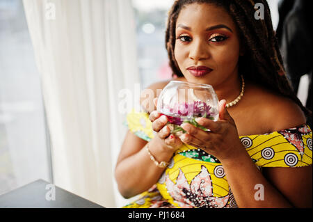 Hauteur petit mignon african american girl avec des dreadlocks, d'usure au niveau de la couleur jaune robe, assis au café avec fleur à verre. Banque D'Images