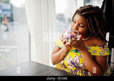Hauteur petit mignon african american girl avec des dreadlocks, d'usure au niveau de la couleur jaune robe, assis au café avec fleur à verre. Banque D'Images