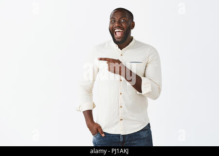 Ha-ha regardez que weirdo. Portrait of happy emotive de bon à l'homme à la peau sombre avec barbe, rire joyeusement, s'amusant sur partie, portant les vêtements à la mode et pointant vers la gauche avec l'index Banque D'Images