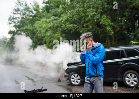 Young man making a phone call après un accident de voiture, la fumée à l'arrière-plan. Banque D'Images