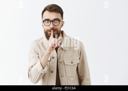 Drôle et mystérieux barbu élégant sous tension avec des lunettes en se baissant veste beige disant chut avec yeux éclaté et l'index sur la bouche de dire plus de secret important mur gris Banque D'Images