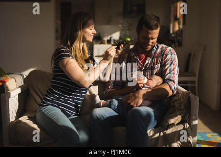 La prise de photo de mère tandis que son père mobile alimentation bébé sur canapé Banque D'Images