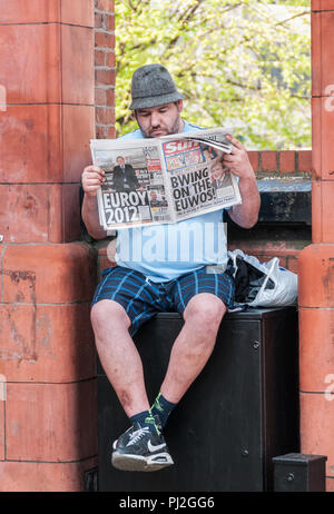 Homme portant un chapeau trilby, short et siégé au mur dans la lecture du journal Le Soleil de Manchester à partir de 2012 avant de l'euro tournoi de football Banque D'Images