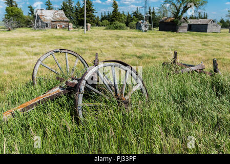 Vieux wagon wheels dans une prairie abandonnée cour avec une vieille ferme, éolienne, et des récipients, dans l'arrière-plan de la Saskatchewan, Canada Banque D'Images