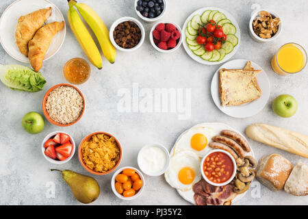 Le petit déjeuner anglais complet, des œufs, saucisses, bacon et champignons avec sélection de fruits et légumes, de pains et de jus sur tableau blanc, gris Banque D'Images