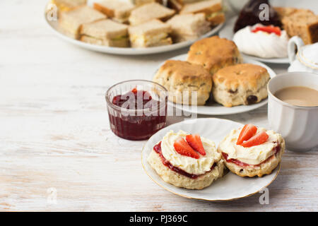 Du thé à la crème anglaise, des scones avec de la confiture et de la crème, thé au lait, avec des sandwichs à l'arrière, sur la table en bois blanc, selective focus copie espace pour t Banque D'Images