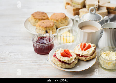 Du thé à la crème anglaise, des scones avec de la confiture et de la crème, thé au lait, avec des sandwichs à l'arrière, sur la table en bois blanc, selective focus copie espace pour t Banque D'Images