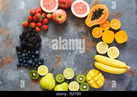 Fruits de couleur arc-en-ciel disposées en cercle, fraises, bleuets, mangue, orange, pamplemousse, banane, pomme, raisins, kiwis, papayes sur le dos gris Banque D'Images