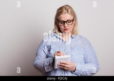 Un sérieux embonpoint femme blonde avec des lunettes et un bloc-notes, de prendre des notes. Banque D'Images
