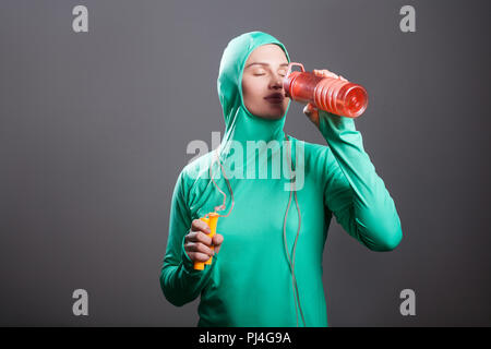 Belle femme musulmane athlète détendue en vert hijab islamique ou sport wear, permanent, de repos holding orange bouteille et de boire avec les yeux fermés. Banque D'Images