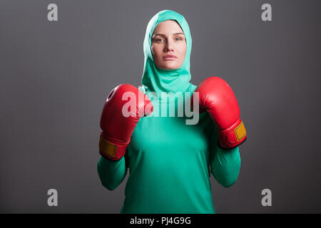 Athlète boxe sérieux femme musulmane en vert hijab islamique ou sport wear debout, avec des gants de boxe et à la caméra avec sérieux à l'intérieur. Banque D'Images