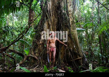 Vieux chaman de la tribu Mentawai couverte de tatoo debout à un vieil arbre dans une jungle dense avec une épée dans ses mains, Siberut, Sumatra, Indonésie Banque D'Images