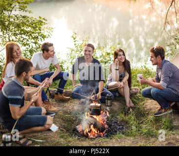 Groupe d'amis sont assis autour d'un feu de camp et de préparer des saucisses. Banque D'Images