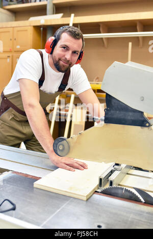 Friendly carpenter avec protège-oreilles et des vêtements de travail travaillant sur une scie dans l'atelier Banque D'Images