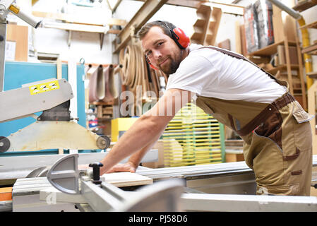 Friendly carpenter avec protège-oreilles et des vêtements de travail travaillant sur une scie dans l'atelier Banque D'Images