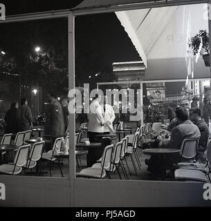 Années 1950, le soir, à Paris, France et deux serveurs chat ensemble sur la chaussée terrasse d'un café sur les Champs-Élysées, juste en bas de l'Arc de Trioumphe monument, le plus grand et le plus célèbre arc dans le monde. Banque D'Images