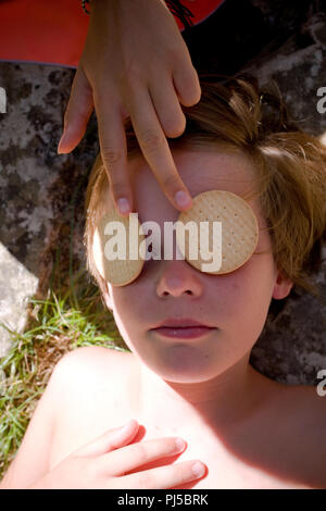 Close up de onze ans la tête et les épaules du garçon allongé à l'ombre d'une main féminine tenant deux biscuits ronds sur ses yeux Banque D'Images