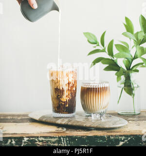 Café glacé dans de grands verres avec du lait versé sur de cruche à la main, mur blanc et de plantes vertes, d'agences à l'arrière-plan, l'espace de copie, square culture. Su Banque D'Images