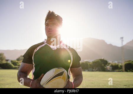 Jeune sportif tenant un ballon de rugby avec un uniforme sale. joueur de rugby sur le terrain avec la lumière du soleil brillante de l'arrière. Banque D'Images