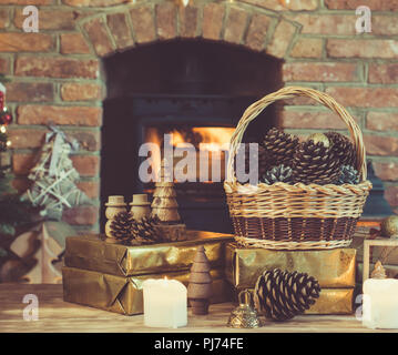 Composition de Noël, des pommes de pin dans panier en osier, objets en bois, présente sur la table en face d'une cheminée avec poêle à bois, bougies et Garland Banque D'Images