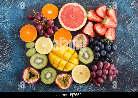 Assiette de fruits : oranges, citrons, pamplemousses, kiwis, fraises, raisins, bleuets, fruit de la passion, la mangue sur l'ardoise noire sur le bleu foncé b Banque D'Images