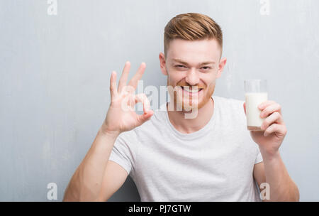 Jeune rousse homme sur mur gris grunge de boire un verre de lait faire ok signe avec les doigts, symbole excellent Banque D'Images