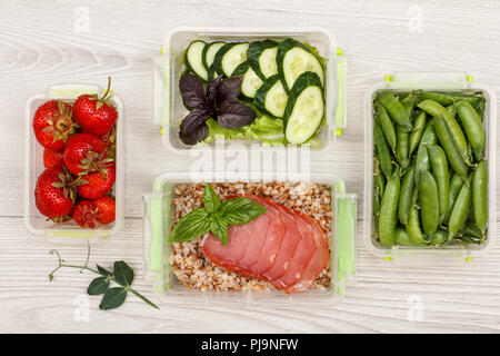Les contenants en plastique préparation des repas avec des fraises fraîches, des pois verts, des concombres et de la salade, à l'eau bouillie de sarrasin et tranches de viande sur fond gris Banque D'Images