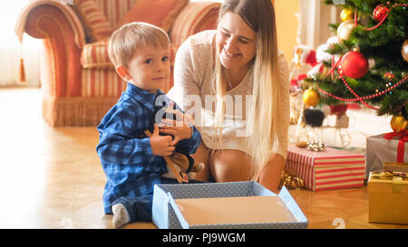 Adorable petit garçon assis sous l'arbre de Noël et serrant peluche il a reçu en cadeau du Père Noël Banque D'Images