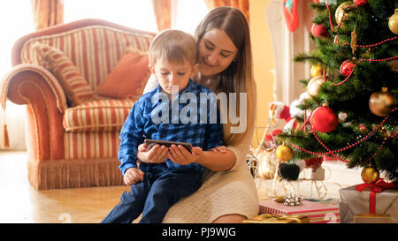 Portrait de jeune maman avec bébé garçon se détendre le matin de Noël et regarder des vidéos sur téléphone mobile Banque D'Images